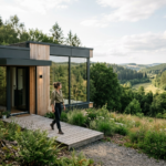 Nowoczesny dom z gotowych elementów z dużymi oknami, otoczony zielenią, z osobą podziwiającą widok.